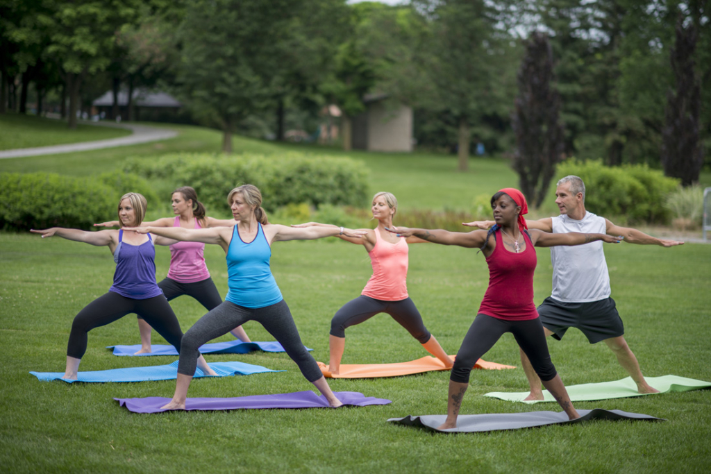 A multi-ethnic group of adults are standing in warrior two pose during their outdoor yoga class at the park.